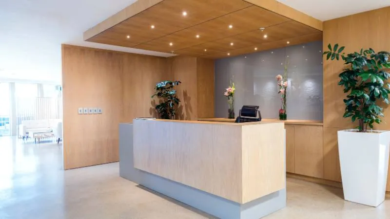 Banco reception in legno chiaro con controsoffitto in doghe e una pianta alta accanto